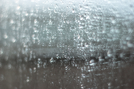 Abstract shiny raindrops on a window glass surface on a gray clouds and dark buildings background. Water drops pattern close up shotの写真素材