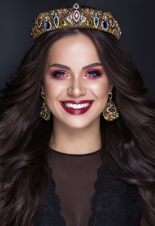 Beautiful brunette girl with a golden crown, earrings and professional evening color make-up and perfect smile. Beauty face. Miss worldの写真素材