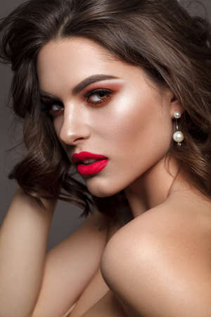 Luxury sexy brunette model with bright professionsl make up, coral eyeshadows, long lashes and red lips. Beautiful wavy hair. Pearl earring. Grey background.の写真素材