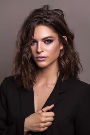 Portrait of young beautiful brunette model with violet professional make up, trendy wavy hairdo and perfect skin. Confident business lady in black fashion jacket on a grey background.の写真素材
