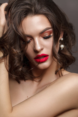Hot sexy brunette model with bright professionsl make up, coral eyeshadows, long lashes and red lips. Beautiful wavy hair. Pearl earring. Grey background.の写真素材