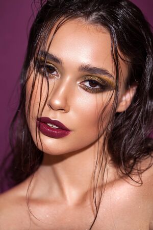 Photo of a beautiful girl with trendy golden eyeshadows, rich eyebrows, marsala lipstick, wet hairdo, violet backgroundの写真素材