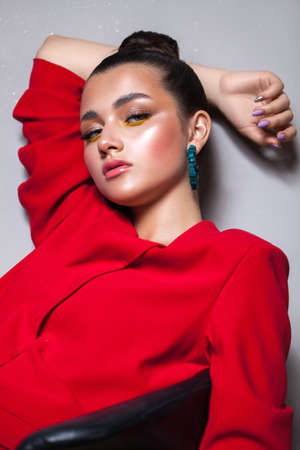 Young beautiful girl with professional make-up, fashionable multi-colored shadows and in a bright red jacket sitting in a relaxed position leaning on the wall. The hair is gathered in a tight, smooth bun, blue-green earrings in the ears.の写真素材