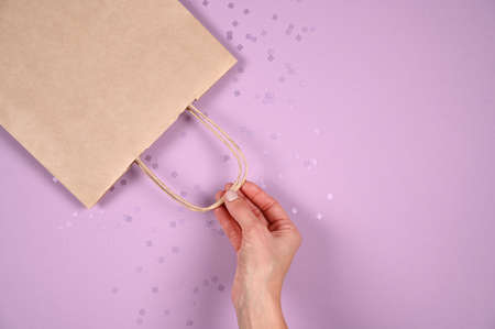 Close up female hold in hand empty craft paper gift bag for purchases after shopping on pastel violet background. Packaging template mockup. Delivery concept advertising mockの写真素材