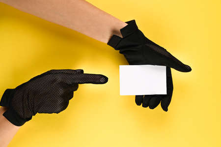 Woman in black gloves holds in one hand a blank visit card with copy space and pointing on it with her index finger of another hand. Contact us.の写真素材