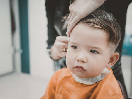 Cute toddler boy on the haircut with a professional childrens hairdresser. Little boy having a stylish haircut at hair salon. Hairdressers hands making hairstyle to child.の写真素材