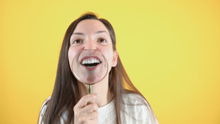 Funny caucasian girl in white t-shirt showing her teeth with magnifying glass, standing over yellow...