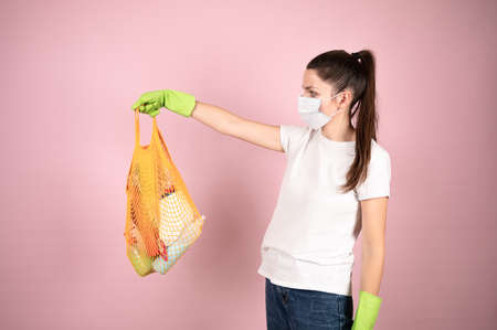 Young brunette in protective mask and gloves holding cleaning supplies in yellow cotton mesh. Zero waste concept. High quality photoの写真素材