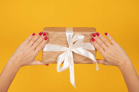 Cropped female hands with perfect red manicure holding wrapped gift box over yellow background. Gift delivery concept. High quality photoの写真素材