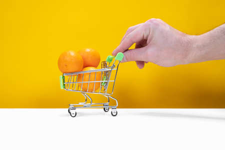 Male hand holding small shopping cart with tangerines on yellow background. Buying fruit concept....