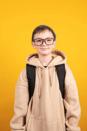 Schoolboy in glasses with bag in biege hoodie. Yellow studio background. Education concept. Hipster style. High quality photoの写真素材