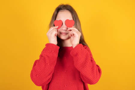 Close-up portrait of cheerful teen girl holding in hands two small little hearts. Young girl hide her eyes isolated on yellow background. High quality photoの写真素材
