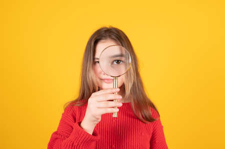 Caucasian schoolgirl looking through the magnifying glass. Teen girl in red sweater on yellow background. Learning concept. High quality photoの写真素材