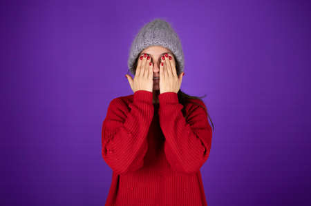 Young woman in warm knitted hat covering her face with her hands. Young woman in red sweater on violet background. High quality photoの写真素材