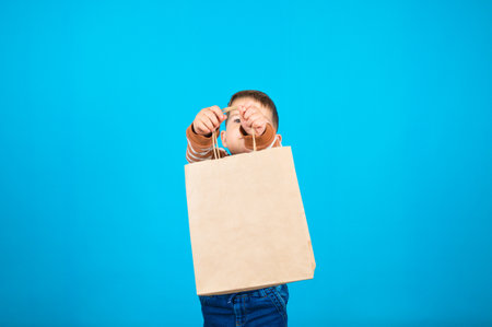 Boy holds out shopping bag isolated on blue. Cute baby boy with paper package with empty space for text or logo. High quality photoの写真素材