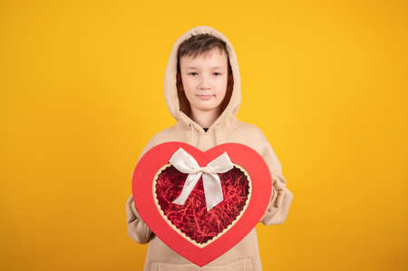 Ten years old boy holding a red heart-shaped gift box in his hand and having fun. Teen boy in biege hoodie on yellow background. Shopping and gift concept. High quality photoの写真素材