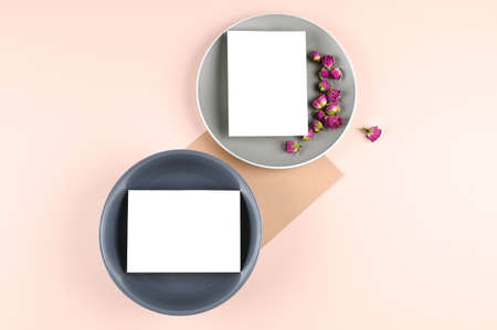 Blank white paper card on gray plate, pink pastel surface with rosebuds. Mock-up of blank greeting card. Minimal business brand template. Flat lay, top view. High quality photoの写真素材