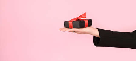 Female hand holding black gift box with red ribbon on pink surface. Sharing, giving or delivery concept. High quality photoの写真素材