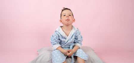 Kid boy in bathrobe sits on pillow and shows his tongue. Child boy refuses to go to bed. High...