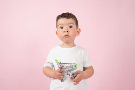 Surprised caucasian boy holds a small shopping trolley on pink background. E-Commerce concept. High...