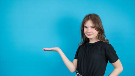 The teen girl in black t-shirt points her palm to the side on blue background, something imaginary...