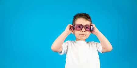 Serious kid boy in white t-shirt wearing violet glasses on blue background with copy space. High...