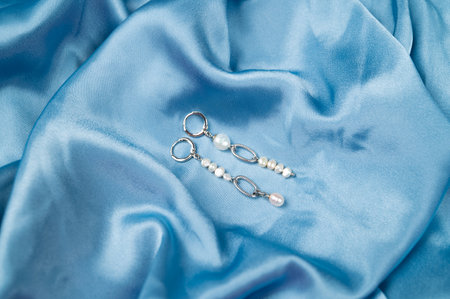 Top view of stylish earrings with pearls on wavy blue satin surface. Fashion and style concept. Top view. High quality photoの写真素材