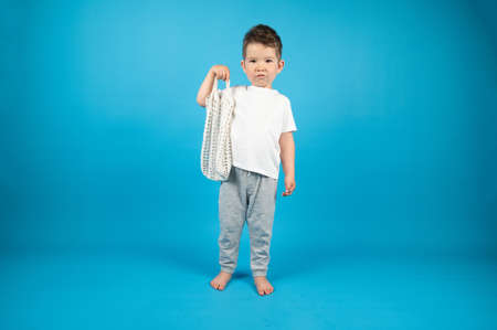 Full-length portrait of serious kid boy with eco friendly mesh bag on blue background. Zero waste...