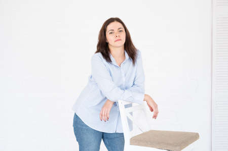 Plus size woman in blue shirt and jeans posing in studio on chair, white background with copy space.の写真素材