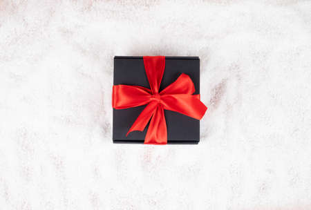 Close-up of black gift box with red ribbon in snow, top view.