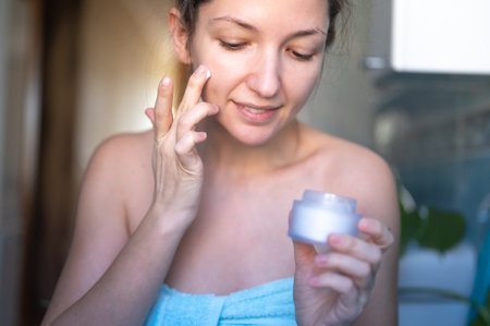 Young beautiful woman applies cream to her face. Skin care at home in the bathroomの写真素材