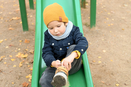 Caucasian toddler boy in a warm clothes is sitting on a childrens slide. A sad independent boy in autumnの写真素材