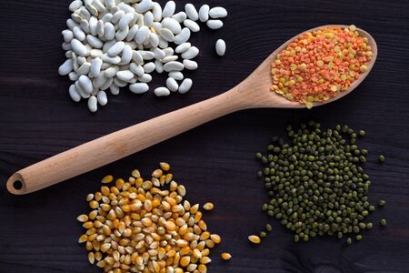 red and yellow lentils, corn, white beans and green mung on a black background. wooden spoon with...