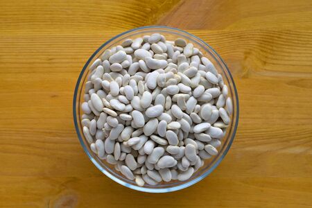bowl of uncooked white beans in a glass bowl on yellow wooden table. top view. Healthy lifestyle.