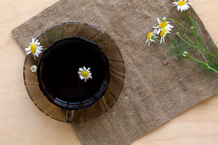 dark glass cup of Herbal tea with chamomile tea on sackcloth on a light wooden table with copy space. cup fo tea with bouquet fresh flowers and green leaves on yellow background. top view.Healthy lifeの写真素材