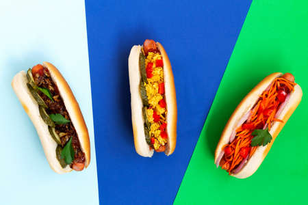 Hot dogs with different toppings: pickles, fried onion, tomatoes and carrots with green on a colour backgrounds with copy space. Fast food concept on the blue, light blue and green.の写真素材