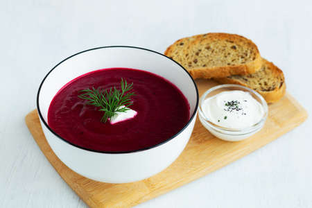 homemade red vegetable soup with greens on the cutting board. white bowl of Beetroot cream soup with...