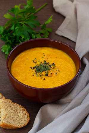 Carrot soup with parsley on brown table. bowl of tasty carrot cream soup with bread and herbs on wooden background. Spring vegetable Homemade cream soup with copy space. verticalの写真素材