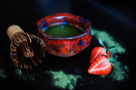 Top-side view of a red chawan of matcha, a chasen and of two halves of strawberry on a black surface with spilled matcha on itの写真素材