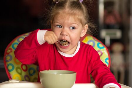 Portrait of little girl eating porridge using spoon sitting in a feeding chairの写真素材