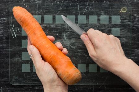 Top view of unpeeled carrot in woman hands with knife on glass cutting board before peeling on black...