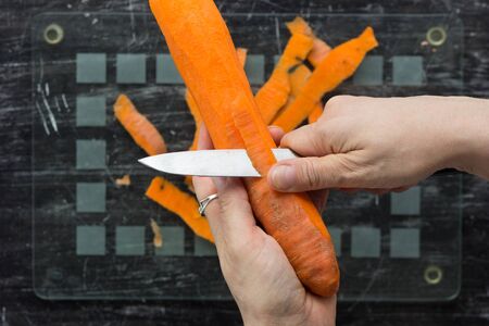 Top view of woman hands peeling carrot with knife on glass cutting board on black backgroundの写真素材