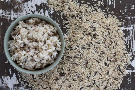 Top view cooked mixed brown and black rice in bowl and heap of raw mixed brown and black rice on the wooden backgroundの写真素材