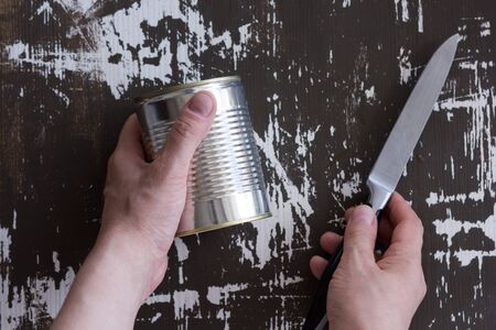 Top view woman hand holding closed can and knife to open it on the wooden background