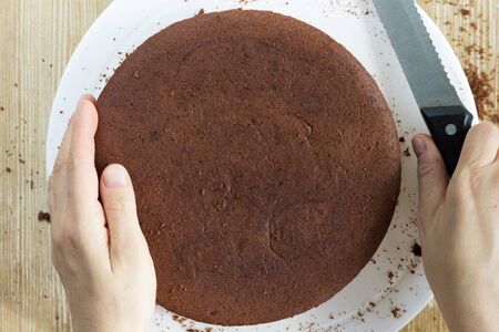 Top view of baked chocolate sponge cake on white plate and woman hands with knife to cut it in half...