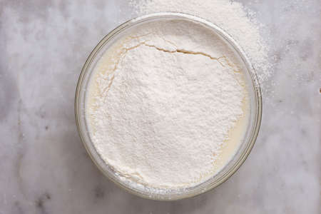 Top view of glass bowl with batter in it covered by sifted flour on marble surface