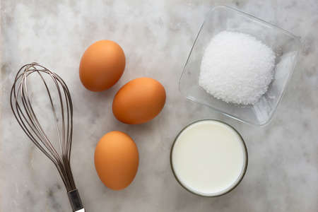 Top view of three raw brown egg, salt, milk and whisk as ingredients of omelette on marble surfaceの写真素材