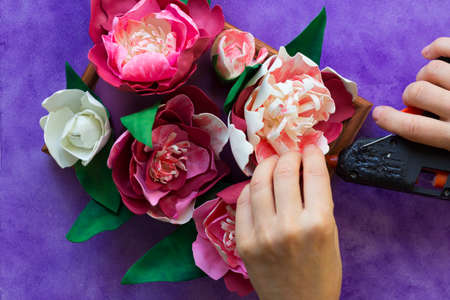 Woman hands glue decorative peony made from foamiran to wooden panel using glue gun on the purple backgroundの写真素材