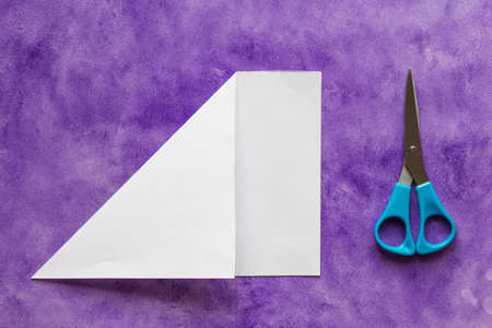 Sheet of paper with folded corner to make square piece and scissors on violet surfaceの写真素材