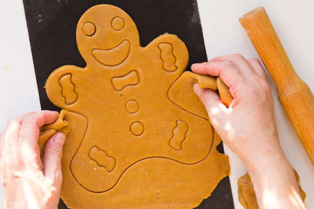 Woman hands removing excess dough after cutting out gingerbread man on raw dough on black baking sheet on white backgroundの写真素材